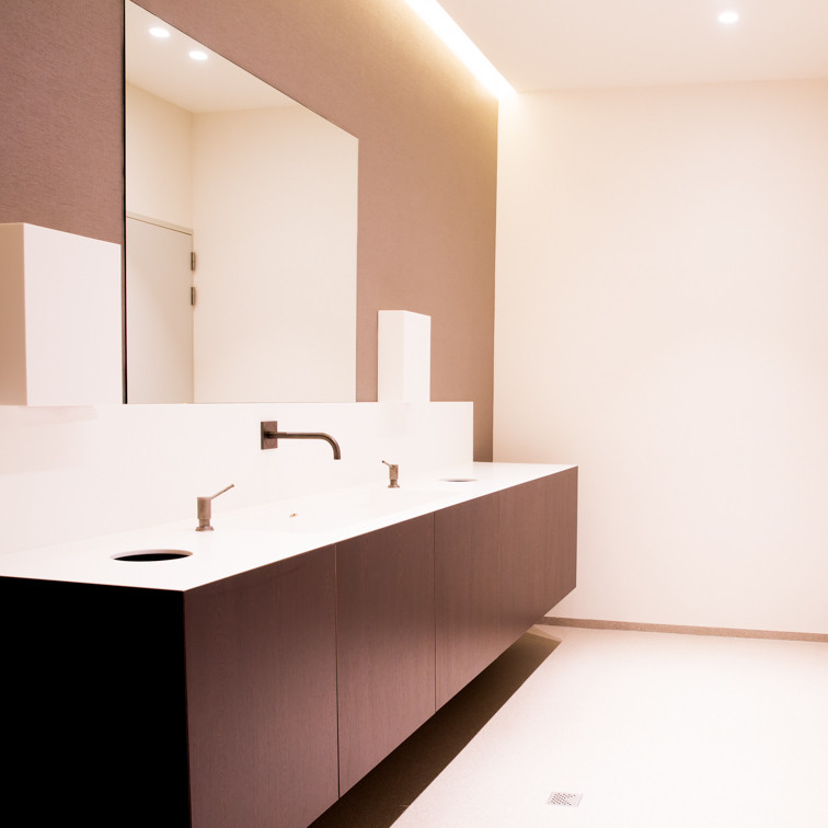 Strakke lijnen, warme materialen en een serene uitstraling - deze badkamer ademt pure rust.<br />De combinatie van het witte werkblad met geïntegreerde wastafels en het donkerhouten meubel zorgt voor een tijdloze, luxueuze look. <br /><br />Het grote spiegelpaneel en de indirecte verlichting versterken het ruimtelijk gevoel en maken van de badkamer een echte oase van elegantie en eenvoud.