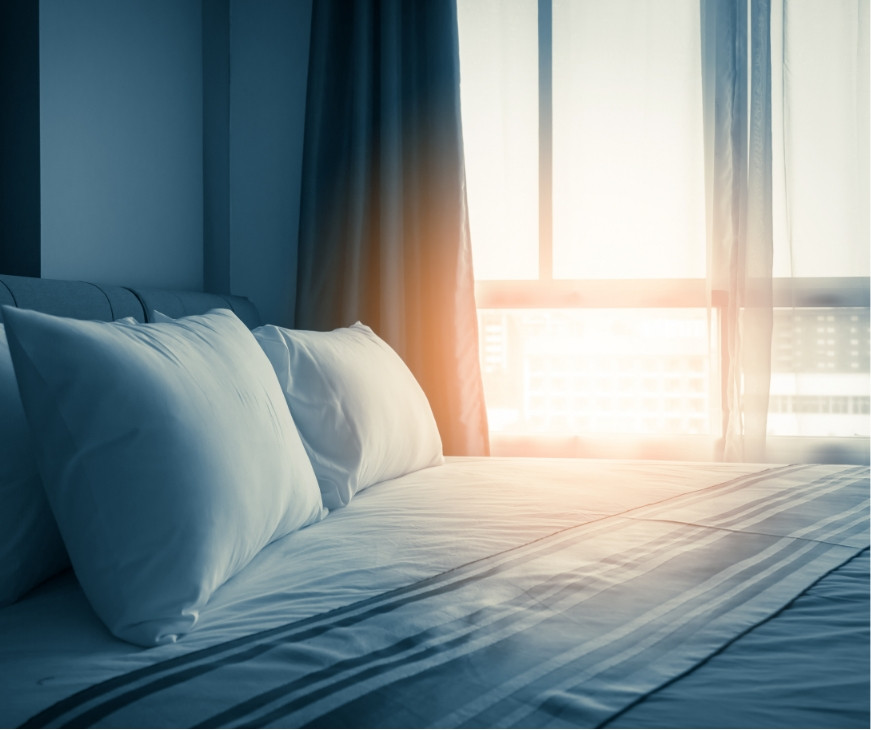 Afbeelding van een opgemaakt bed naast raam met zonlicht